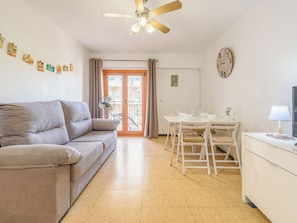 Un salon avec un canapé, une table à manger avec des chaises, un ventilateur de plafond et un coin cuisine équipé d’un réfrigérateur.
