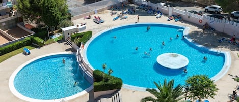A large outdoor swimming pool with multiple sections, surrounded by lounge chairs and palm trees.