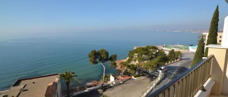 A balcony with a view of the sea, a road, and a building with a white facade.