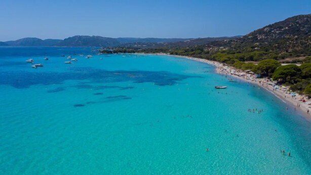 Une plage côtière aux eaux turquoise et limpides, avec des bateaux et une rive sablonneuse où se promènent des gens.