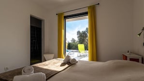 Une chambre d’hôtel avec un lit, une fenêtre avec des rideaux jaunes, une vue sur une piscine et une chaise à l’extérieur, ainsi qu’une petite table avec une lampe.