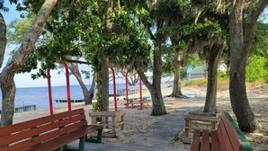Una terraza de madera con bancos y mesas de picnic, rodeada de árboles y con vista a una playa.