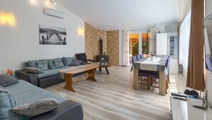 Ein modernes Wohnzimmer mit einer Couch, einem Couchtisch und einem Essbereich mit Tisch und Stühlen. Ein Kamin und ein großes Fenster mit Vorhängen sind ebenfalls vorhanden.