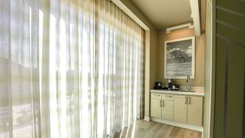 A hotel room with a large window covered by sheer curtains, a wooden floor, a small kitchenette with a coffee maker, and a framed picture on the wall.