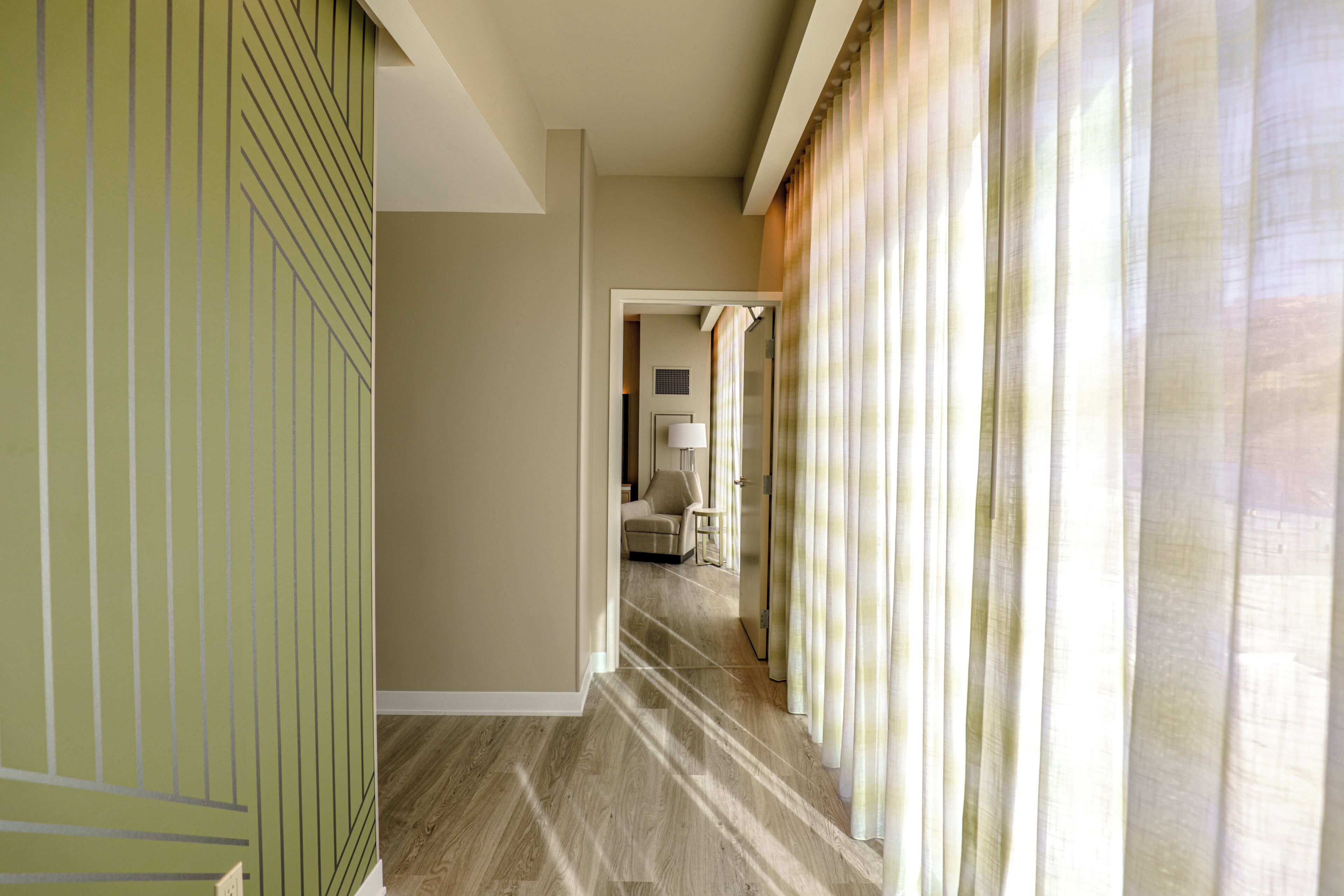 A hotel corridor with a wooden floor, beige walls, and a view into a room with a bed and a lamp.