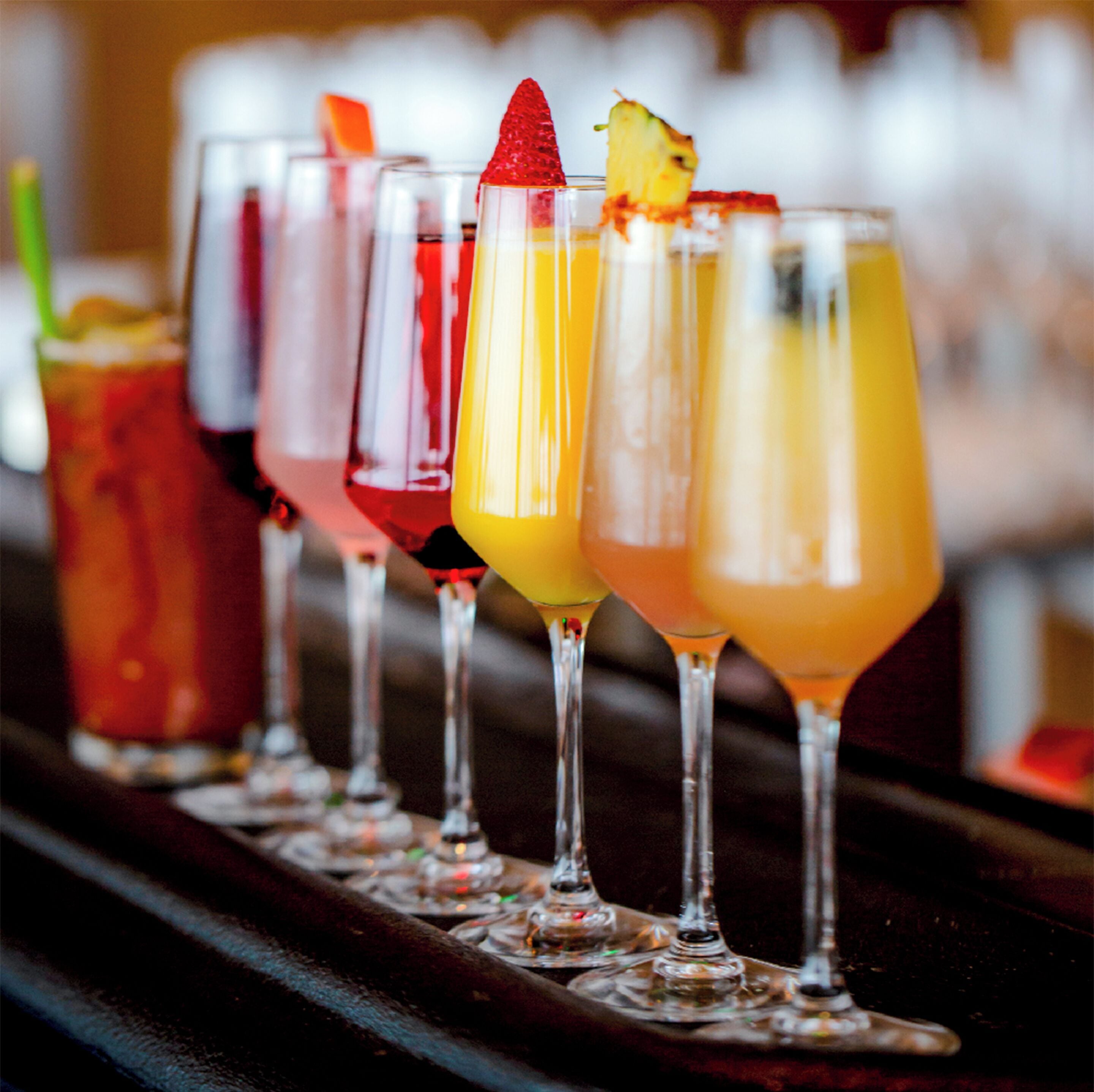 A row of colorful cocktails in glasses with garnishes, displayed on a bar counter.