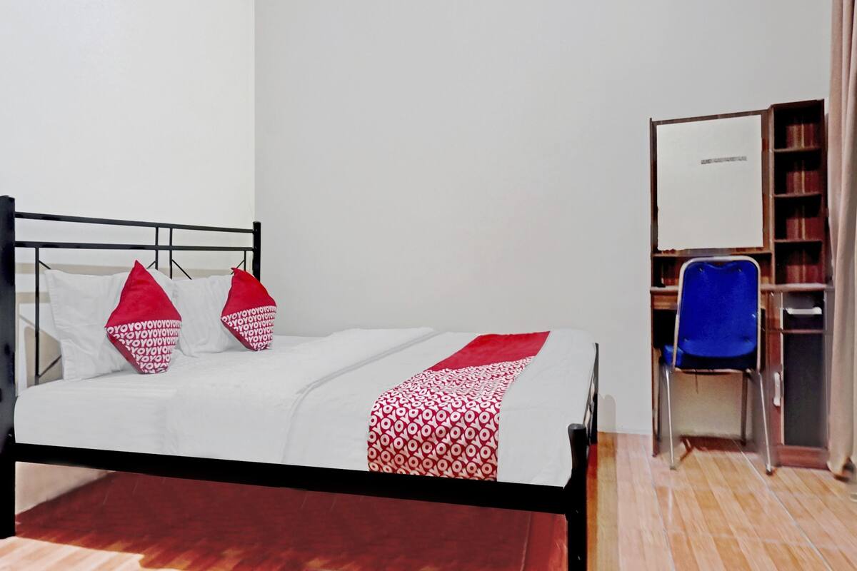Una cama con colcha blanca y roja estampada, dos almohadas rojas y un marco de metal negro. A la derecha, una silla azul y una estantería llena de libros.