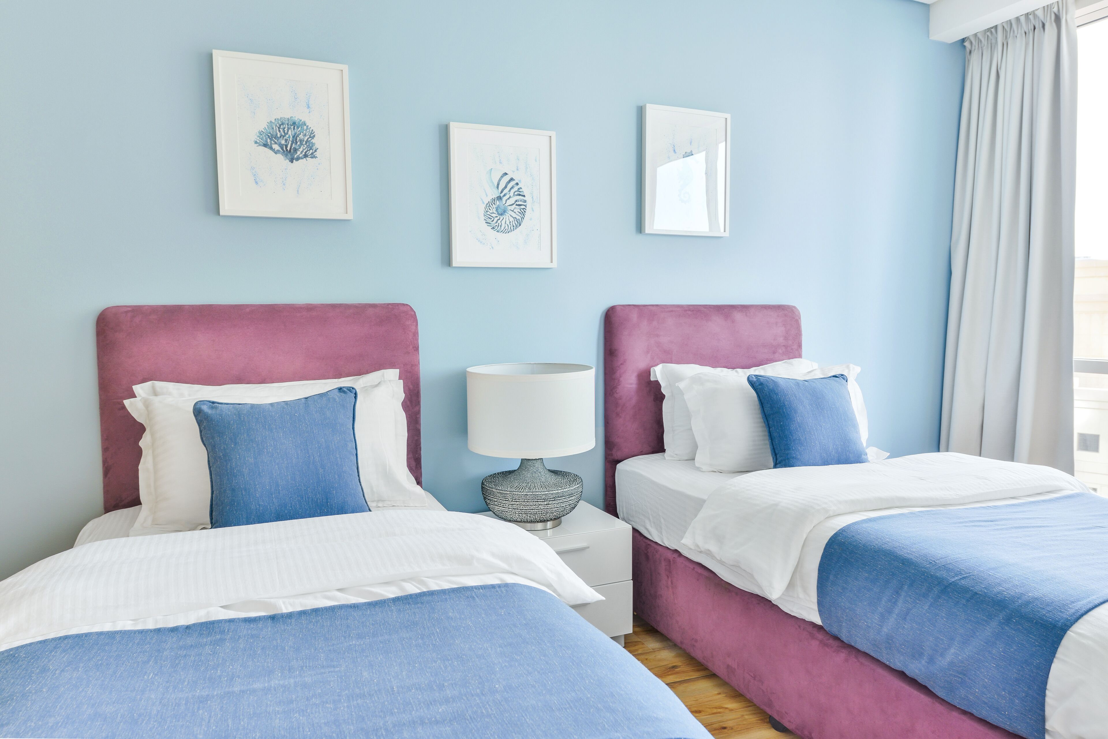 A hotel room with two beds, each with a pink headboard and blue bedding. There are three framed artworks on the wall, a bedside table with a lamp, and a window with curtains.