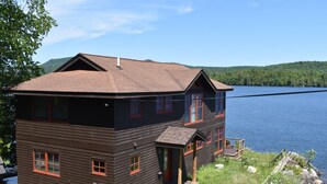 A two-story cabin with a dark wooden exterior and red trim, situated by a lake with a view of green hills in the background. - Stunning Blue Mountain Lake - Waterfront Property! (Blue Mountain lake)
