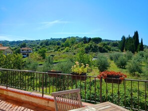 A balcony with a wooden bench and potted plants, overlooking a lush green landscape with trees and a hill in the background.
