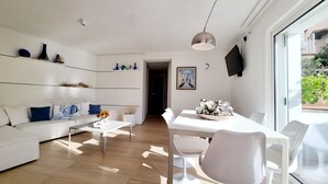 Ein modernes Wohnzimmer mit einem weißen Sofa, einem Couchtisch und einem Essbereich mit weißem Tisch und Stühlen. Ein großes Fenster lässt natürliches Licht herein und bietet einen Blick ins Grüne.