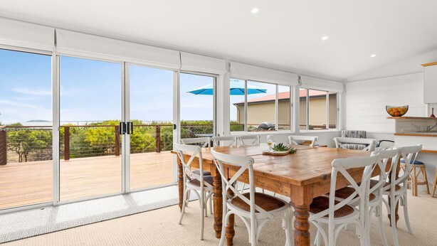 A spacious dining area with a wooden table and white chairs, large sliding glass doors leading to an outdoor deck, and a view of the ocean. - SilverTide Retreat in Silverleaves (Silverleaves)