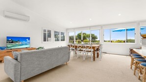 A spacious living area with a grey sofa, wooden dining table, and chairs, large windows with blinds, and a flat-screen TV displaying a tropical beach scene. - SilverTide Retreat in Silverleaves (Silverleaves)