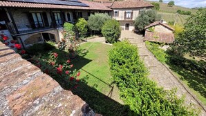 Un bâtiment en pierre avec un toit de tuiles, un jardin parsemé de fleurs rouges et, au loin, un vignoble.