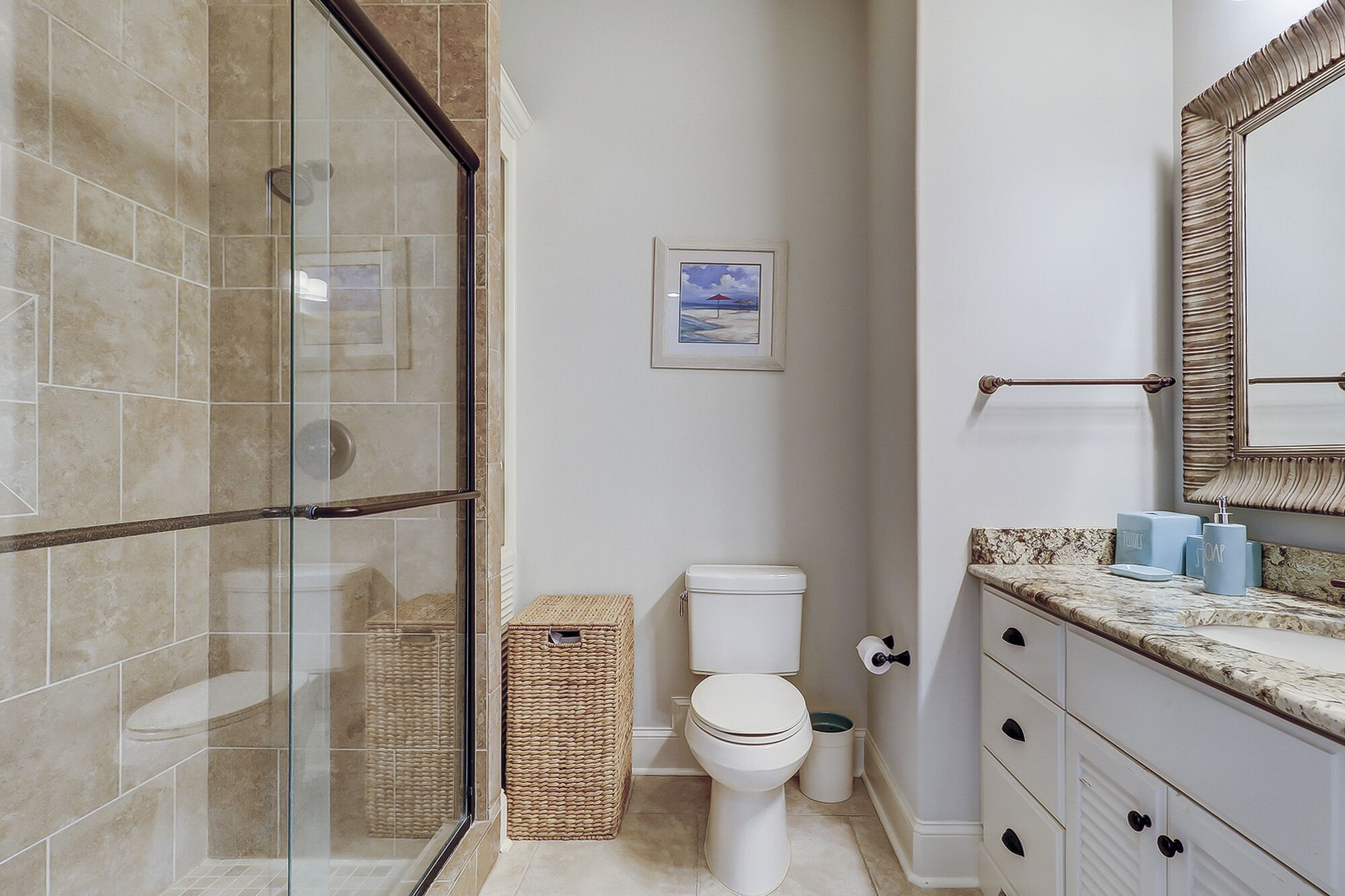 A bathroom with a glass-enclosed shower, a white toilet, a framed picture on the wall, and a vanity with a mirror and toiletries.