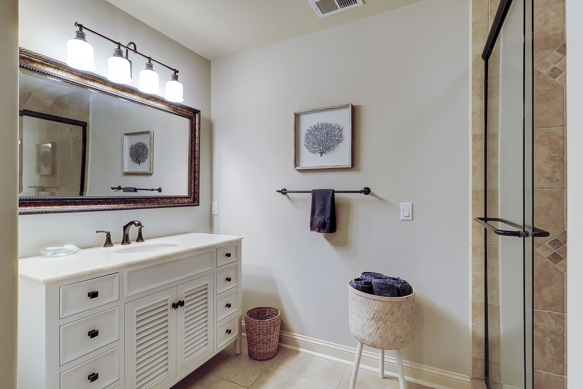A bathroom with a white vanity, a large mirror, a framed tree artwork, a towel rack, and a shower area.