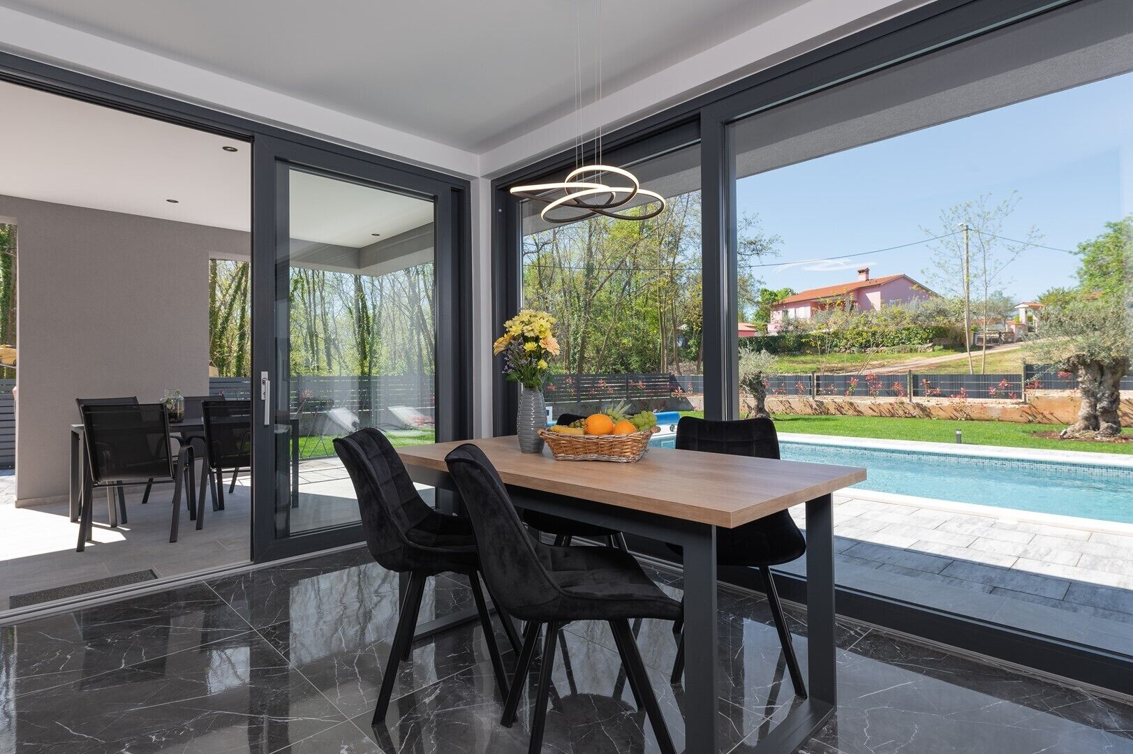 Un comedor moderno con suelo de mármol, sillas negras y una mesa de madera con una cesta de frutas.