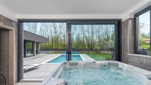 Área moderna de piscina interna com ofurô, grandes portas de vidro deslizantes e vista para um quintal com piscina e vegetação.