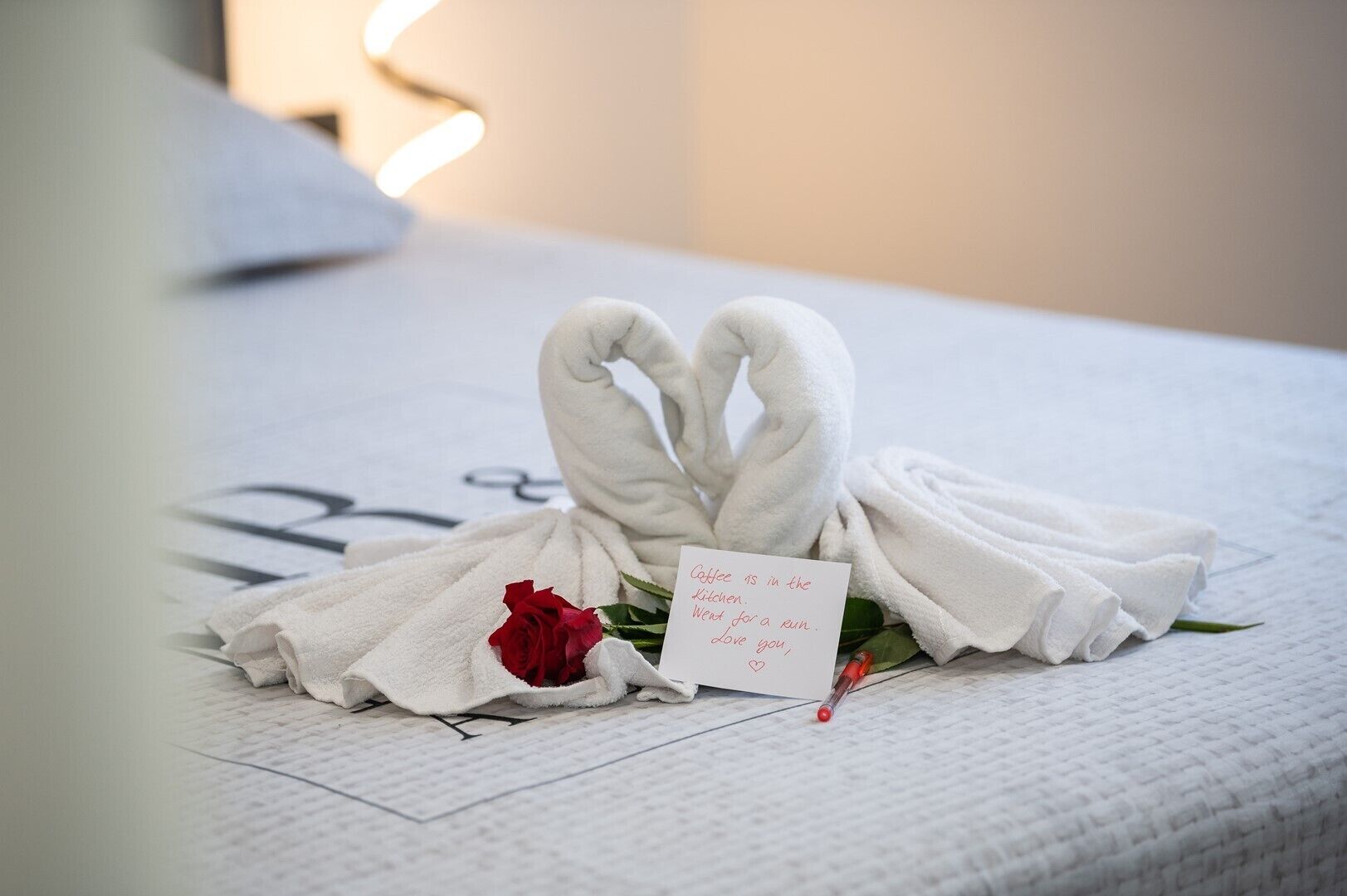 Een keurig opgemaakt bed met handdoeken gerangschikt in de vorm van een hart, een rode roos en een briefje op het bed.