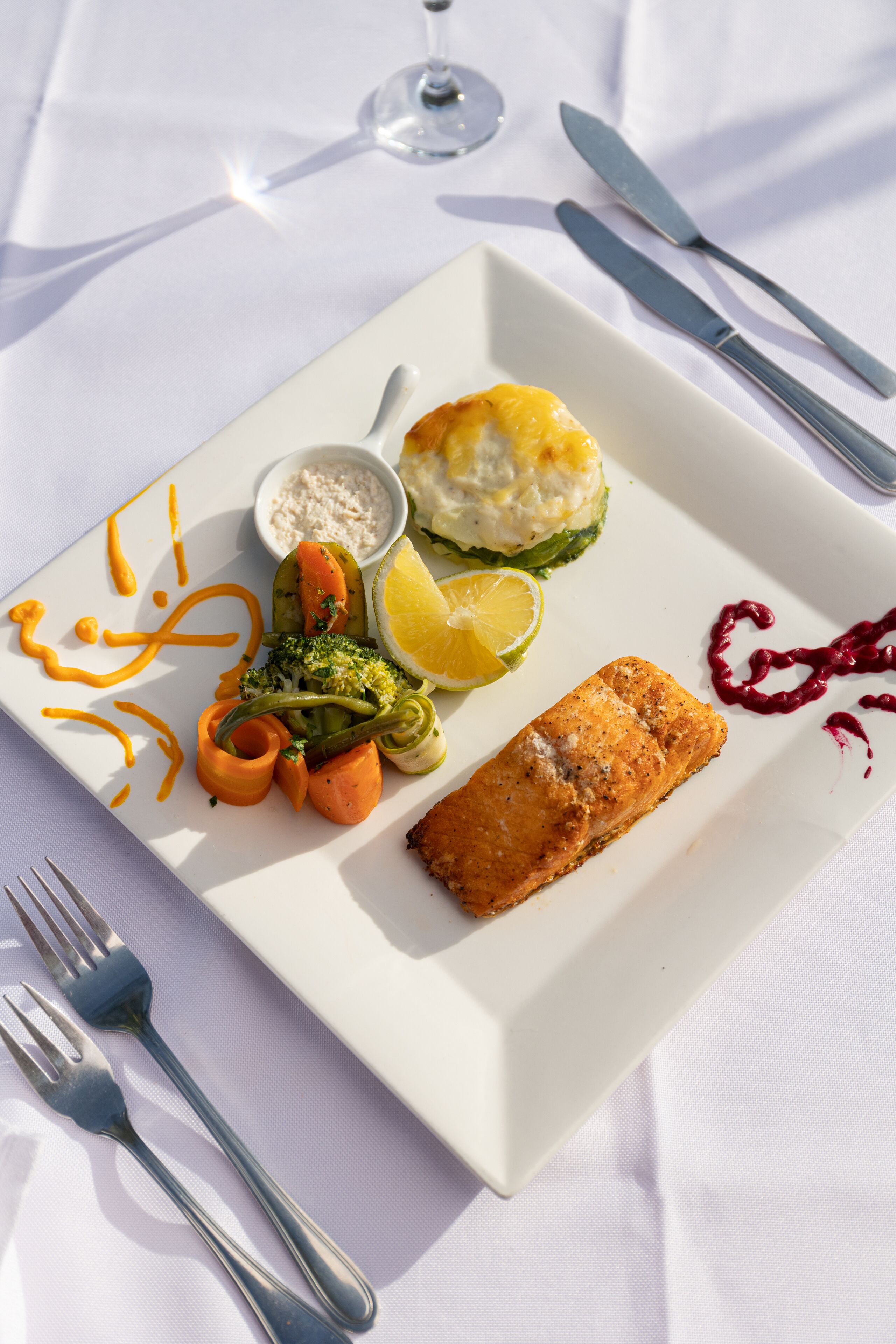 Un plato blanco rectangular con un pescado empanizado, verduras y un recipiente pequeño con salsa.