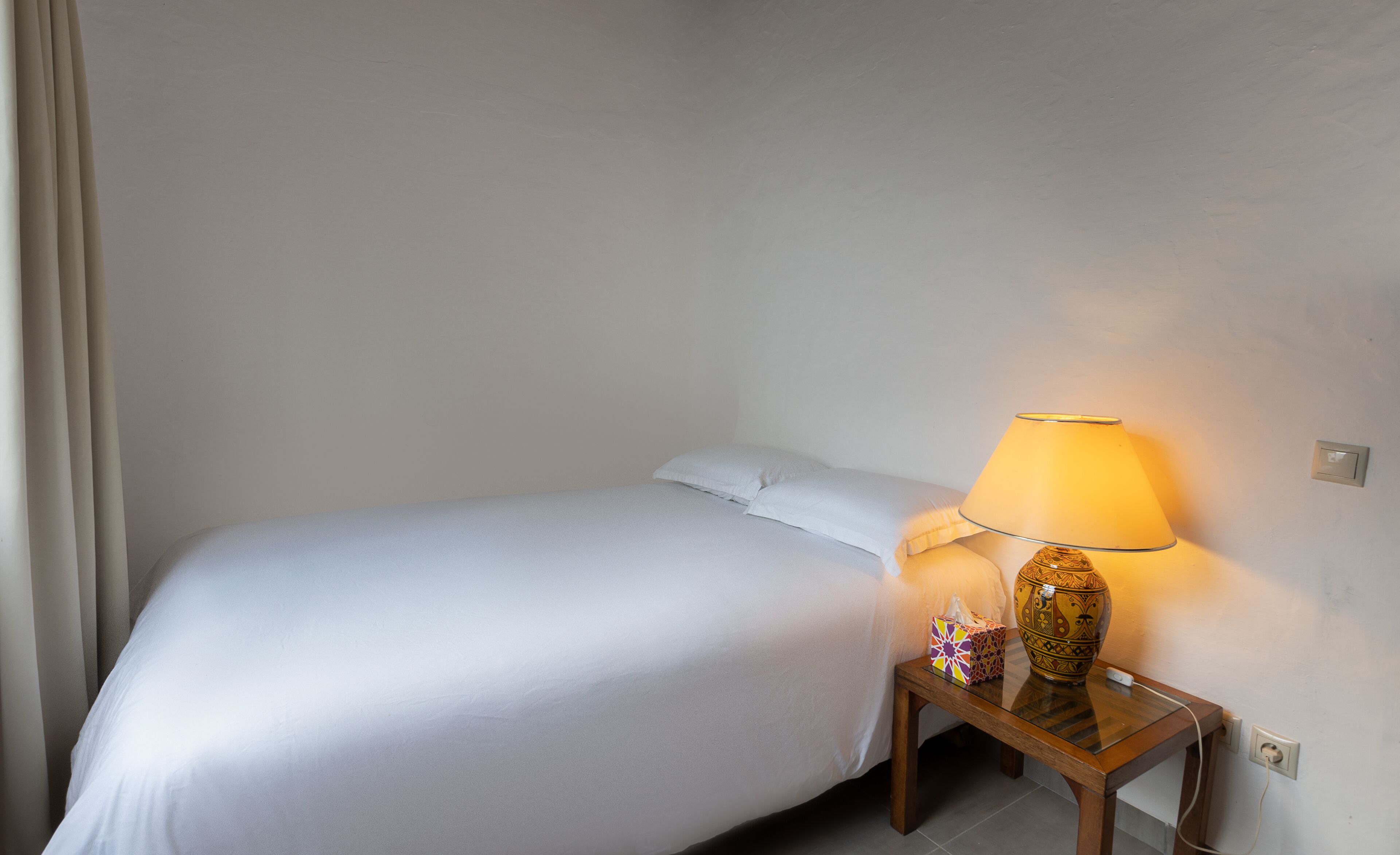 Un lit bien fait, avec du linge de lit blanc, une table de chevet avec une lampe et un petit objet décoratif.
