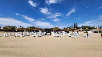 Une plage bordée de rangées de chaises longues blanches et d’ombrelles en chaume, sous un ciel bleu dégagé.