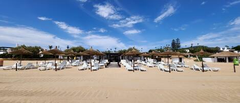 A beach with rows of white lounge chairs and thatched umbrellas, under a clear blue sky.