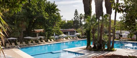 Piscina con sillas de descanso y sombrillas, rodeada de vegetación tropical y un edificio con techo de tejas.