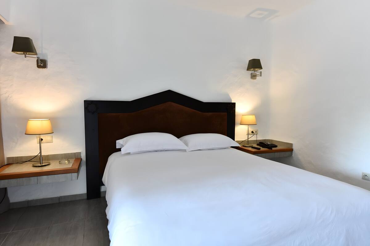 A neatly made bed with white linens, two bedside lamps, and a wooden nightstand with a phone and remote control.