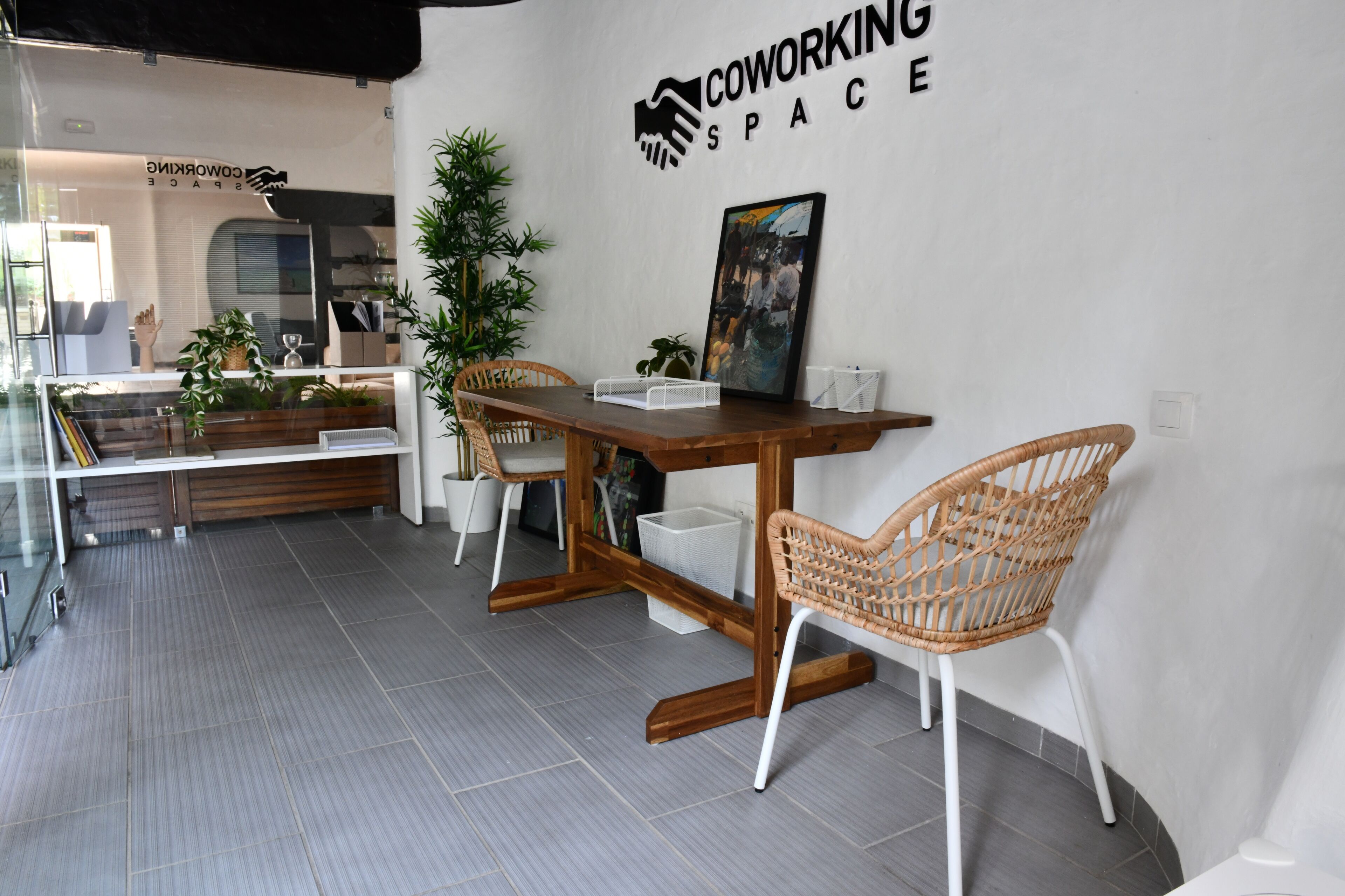 Un espacio de coworking con una mesa de madera, sillas de mimbre, un cubo de basura blanco y un cuadro en la pared.