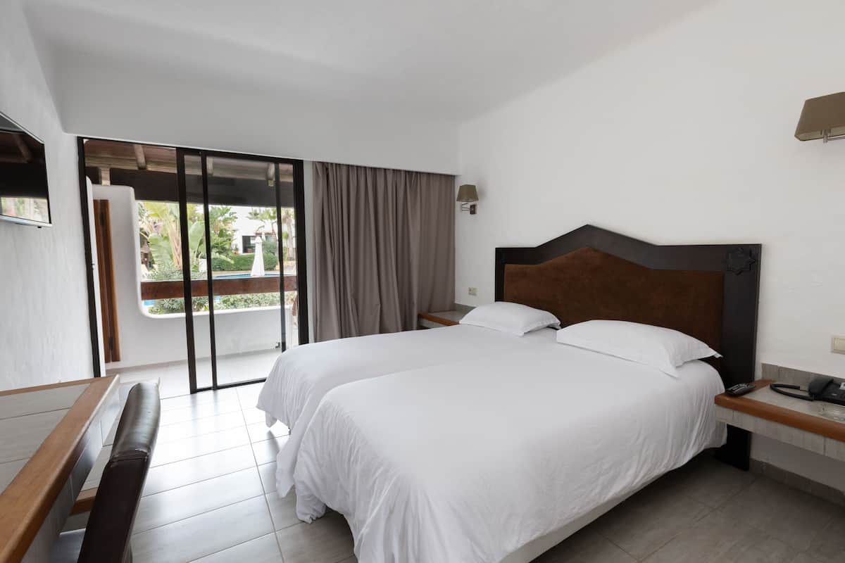 A hotel room with two beds, a bench, a desk, and a sliding glass door to the balcony.