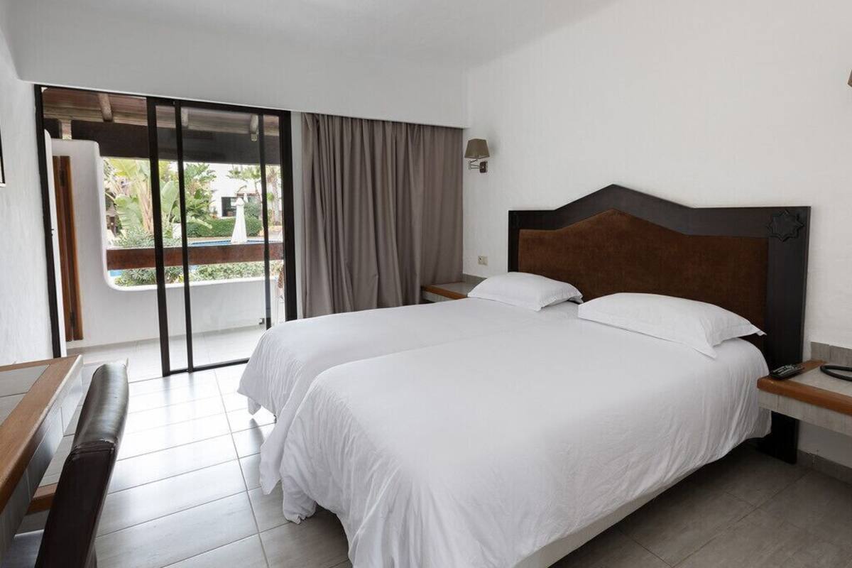 A hotel room with two beds, a bench, a desk, a nightstand, a lamp, and a view of the outdoors.