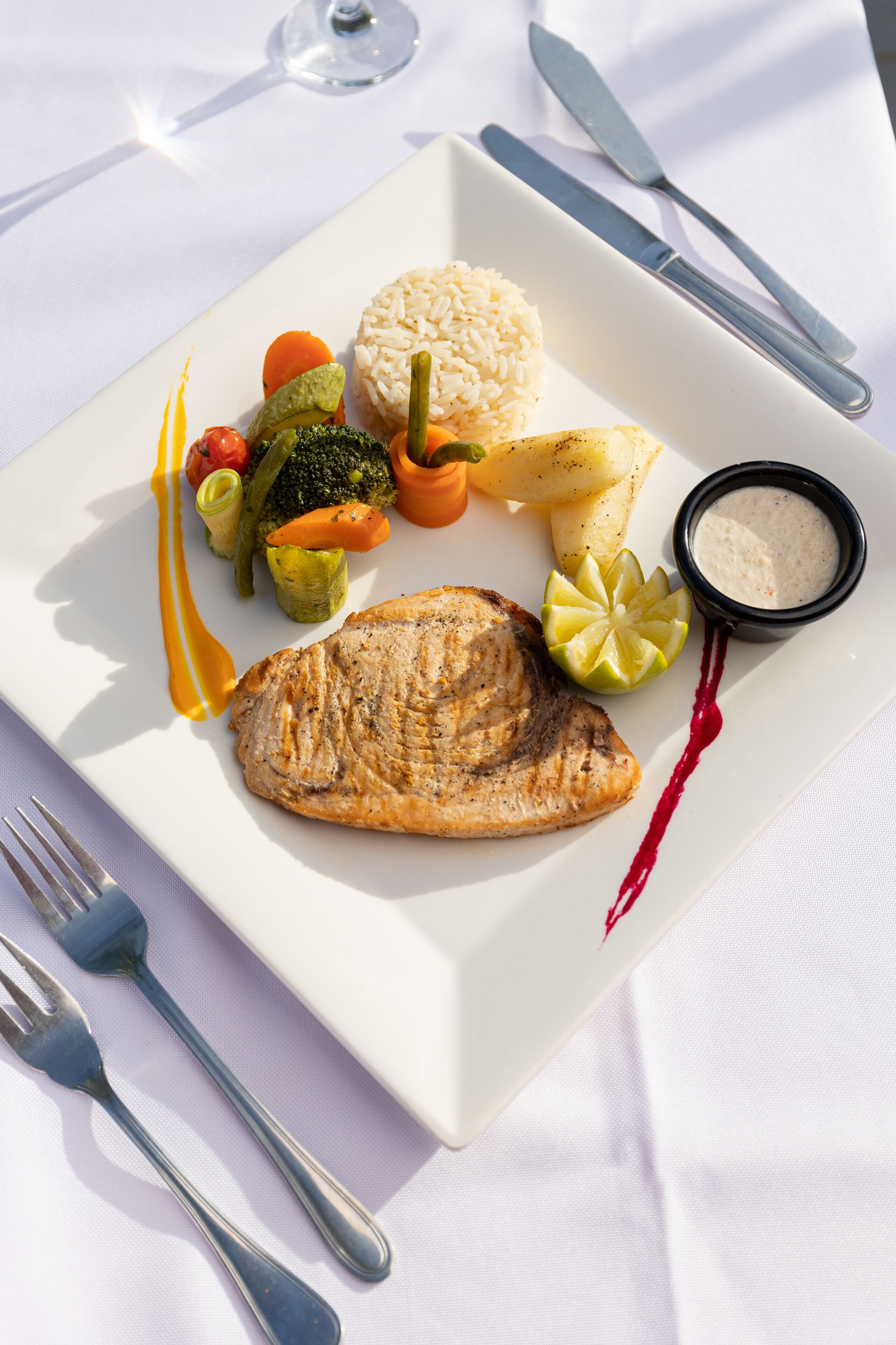 A square plate with a grilled fish, rice, and assorted vegetables, accompanied by a small bowl of sauce and a lemon wedge.