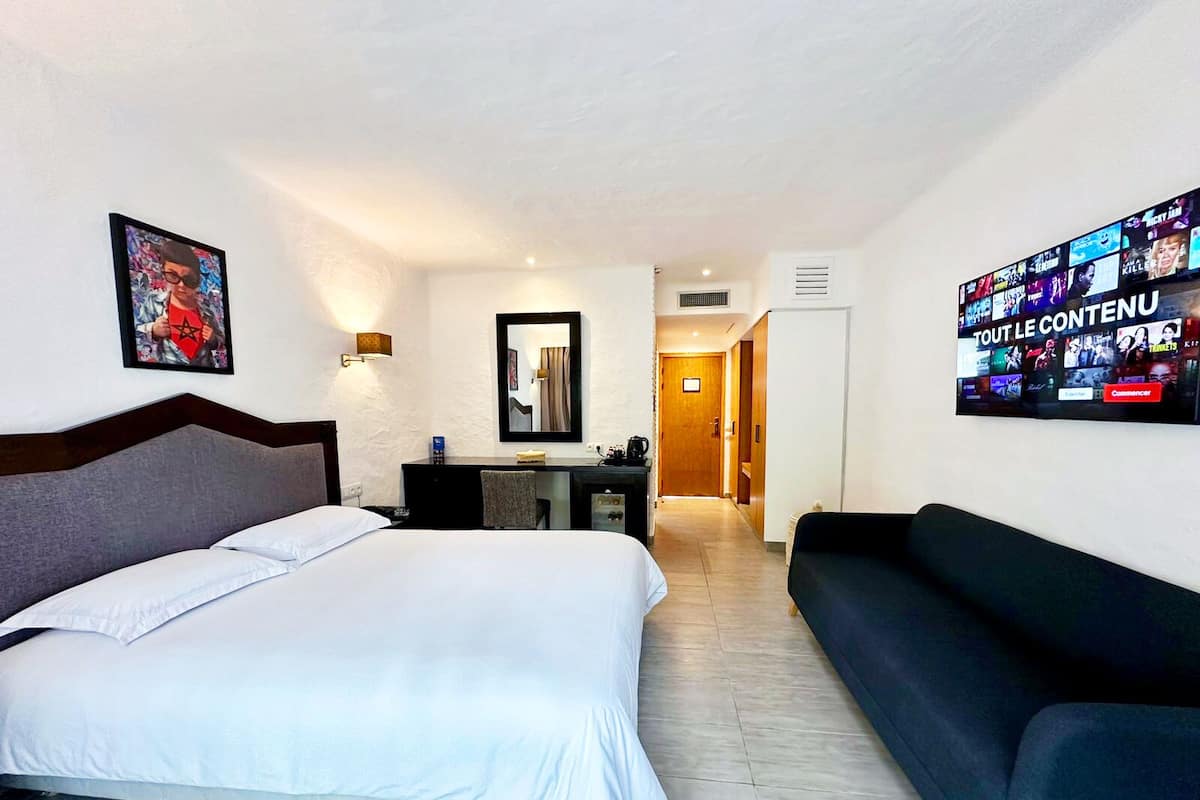 A hotel room with a bed, a desk, a chair, a couch, a TV, and a framed picture on the wall.