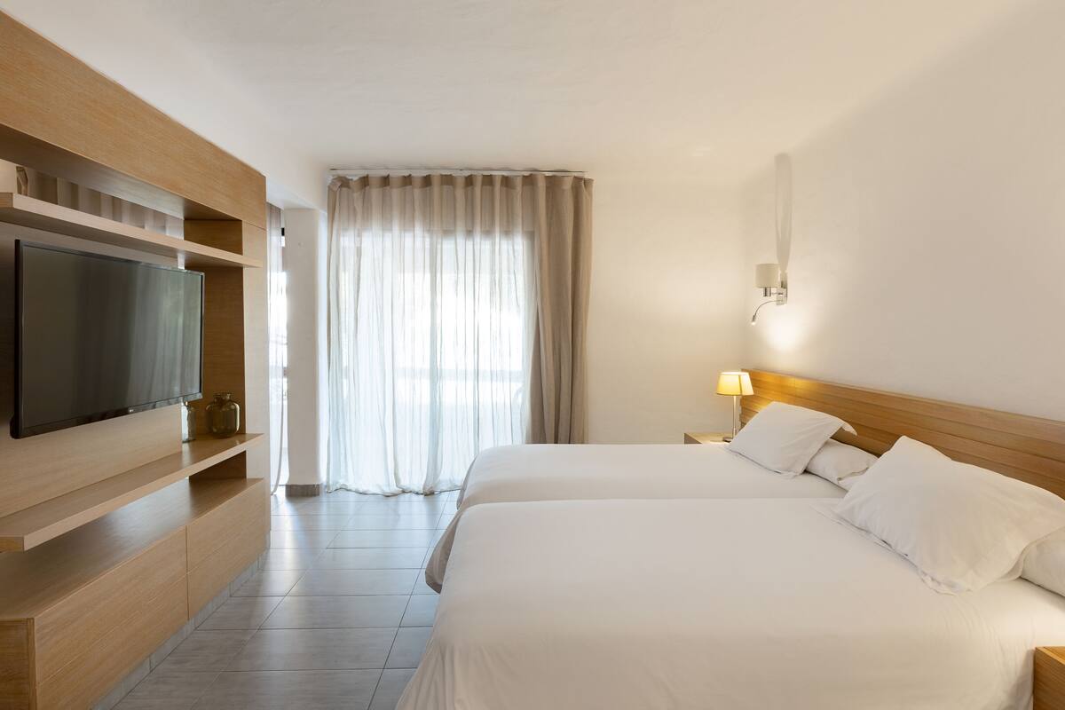 A hotel room with a large flat-screen TV mounted on a wooden shelf, two beds with white linens, a bedside table with a lamp, and a large window with sheer curtains.