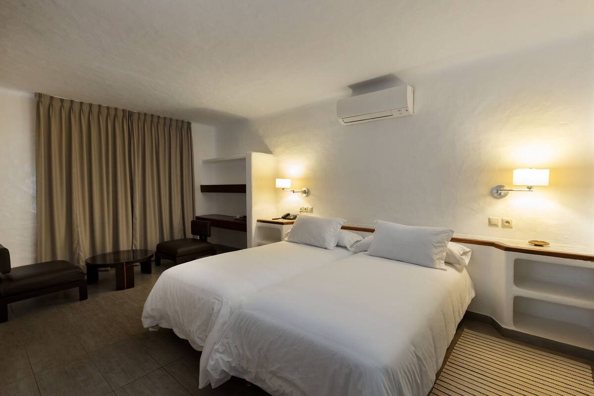 A hotel room with a large bed, two bedside tables with lamps, a desk, a chair, and a large window with curtains.
