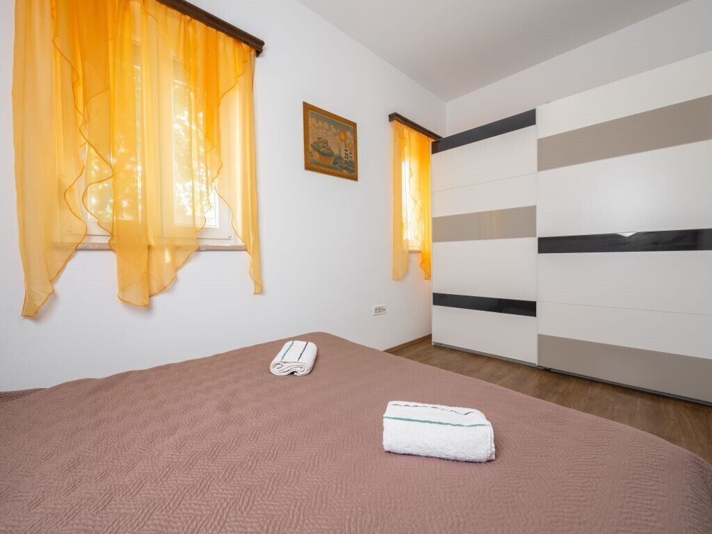 Une chambre d’hôtel avec un lit, une fenêtre ornée de rideaux jaunes, un tableau accroché au mur et une armoire à rayures horizontales.