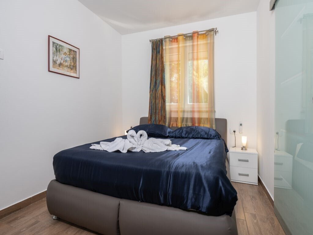 A neatly made bed with a dark blue comforter and pillows, a white bedside table with a lamp, and a framed picture on the wall.