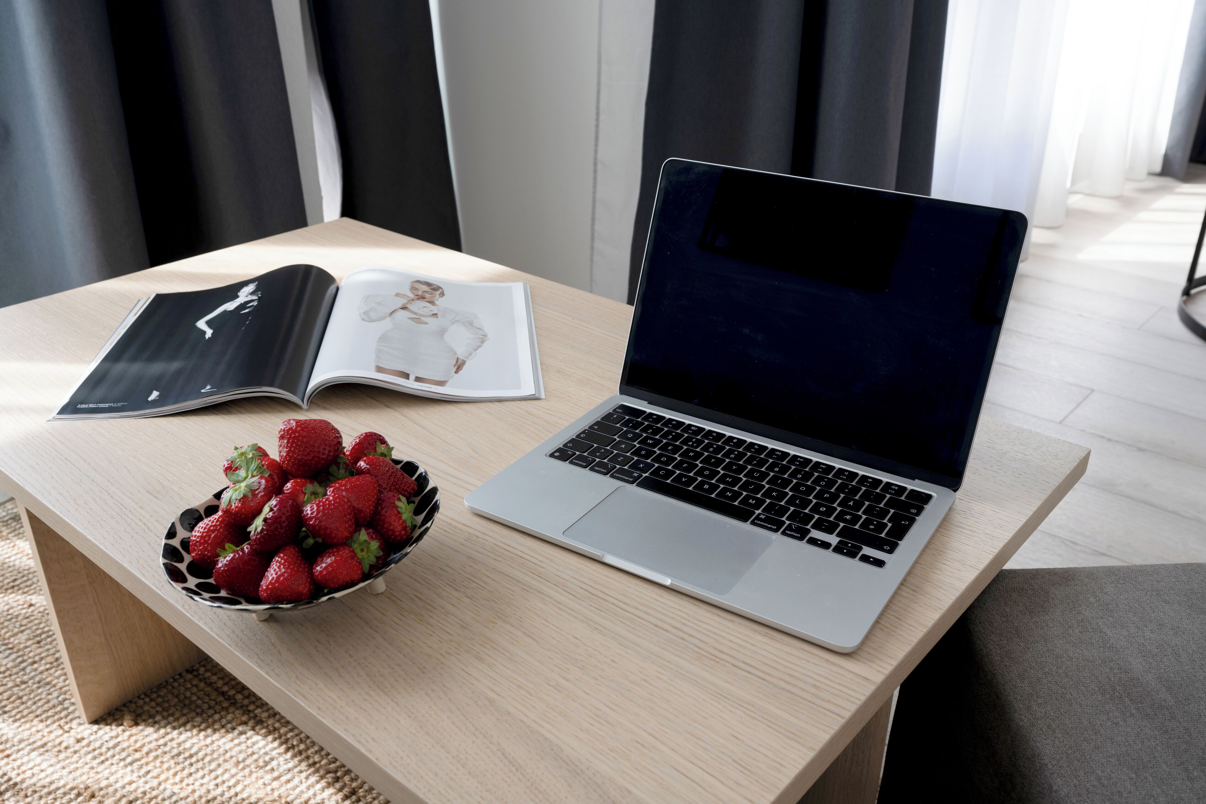 A laptop on a wooden table with a bowl of strawberries and an open magazine.