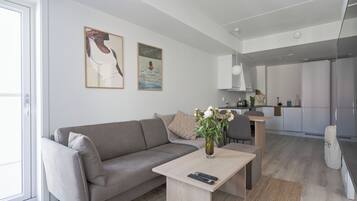 Una sala de estar moderna con un sofá, una mesa de centro con un jarrón, y una cocina al fondo.