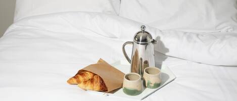 A neatly made bed with white linens, a silver coffee pot, two small cups, and a croissant on a tray.