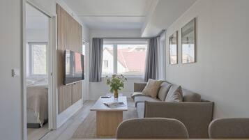 A modern living room with a sofa, coffee table, and TV. There is a window with curtains and a view of buildings outside.