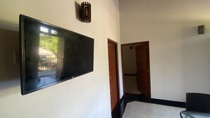 A room with a flat-screen TV mounted on the wall, a wooden door, a glass table, and a view of the outside. - The Kingslayer Resort  (Negombo)