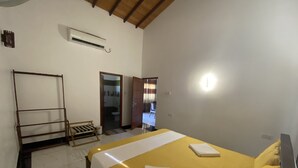 A bedroom with a yellow bed, a wooden bench, a white air conditioning unit, and a door leading to a bathroom. - The Kingslayer Resort  (Negombo)