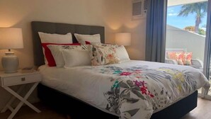 A neatly made bed with a floral bedspread, a bedside table with a lamp, and a view of the outdoors through a window.