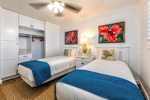 A hotel room with two beds, a wardrobe, a ceiling fan, and two wall-mounted paintings. - Menehune Shores #220 – Beachside Bliss with Panoramic Ocean Views (Kihei, Maui)