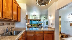 A kitchen with wooden cabinets, a stainless steel sink, a marble countertop, a ceiling fan, and a view of the outdoors. - Menehune Shores #220 – Beachside Bliss with Panoramic Ocean Views (Kihei, Maui)