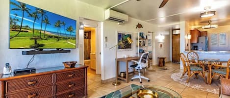 Una sala de estar con televisor, un comedor con mesa y sillas, un ventilador de techo y una cocina.