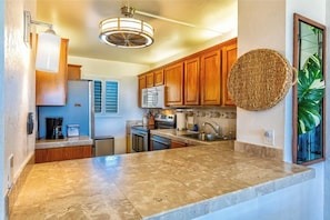 A kitchen with wooden cabinets, a granite countertop, a microwave, a stove, and a hanging woven basket. - Menehune Shores #220 – Beachside Bliss with Panoramic Ocean Views (Kihei, Maui)