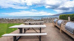 En udendørs terrasse med picnicborde og grill, med udsigt over en stenet kystlinje og havet.
